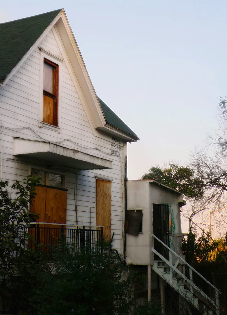 White house with boarded windows and a deteriorated side staircase amid vegetation.