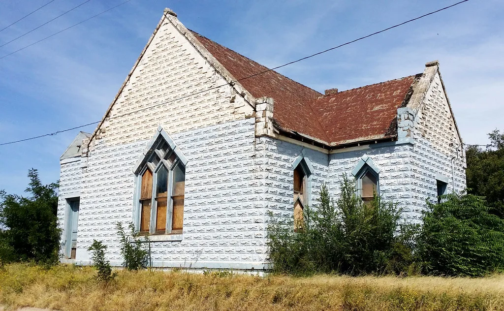 Ancienne église blanche aux fenêtres condamnées et aux abords envahis.