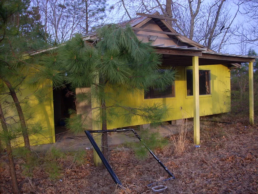 Yellow house with a torn roof and open doorway, with a fallen window frame before pine trees.