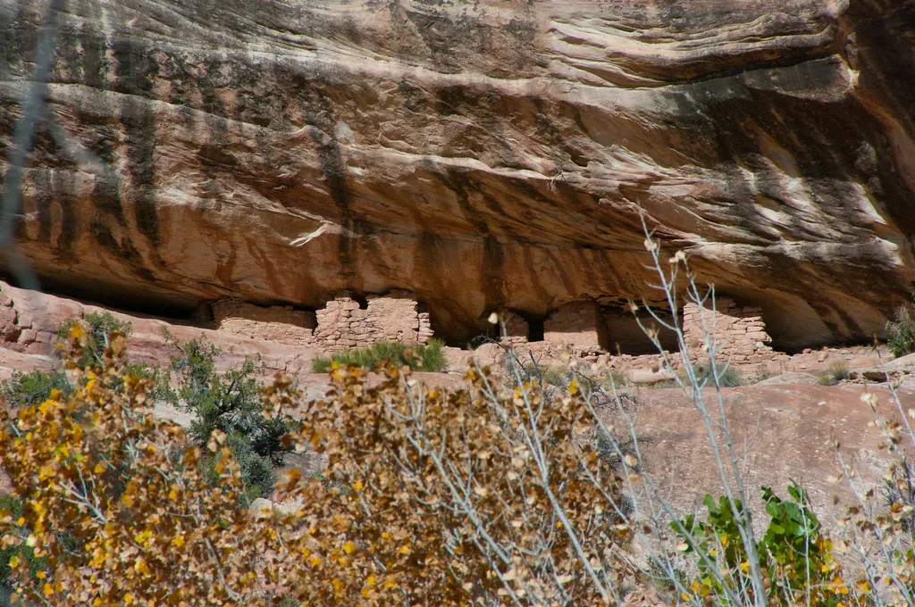 Ruins of small stone buildings beneath a large overhanging rock face.