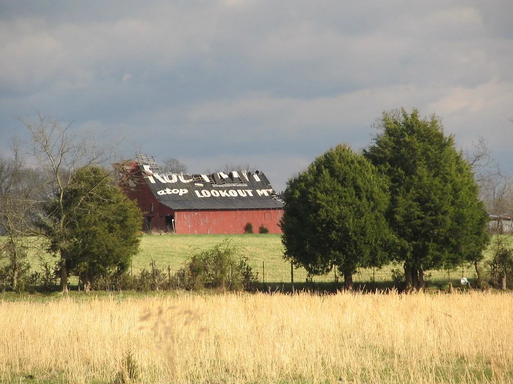 Old red barn with a partly torn-off roof behind trees.