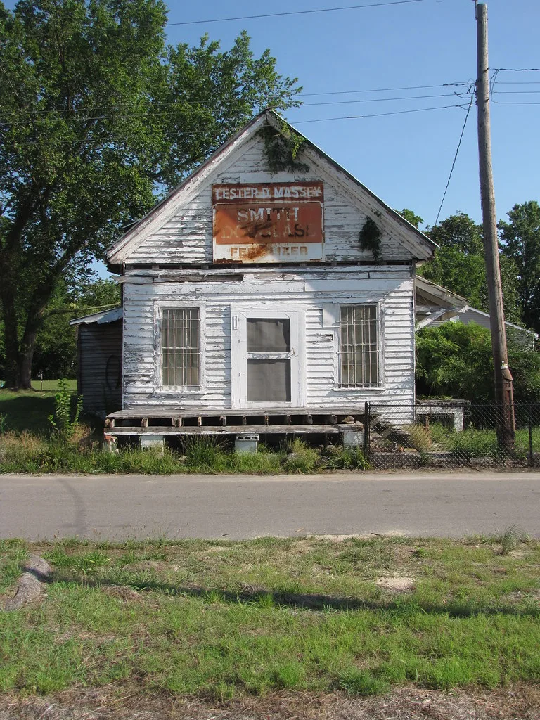 Ancien petit commerce blanc en bois au porche dégradé et à l’enseigne effacée.