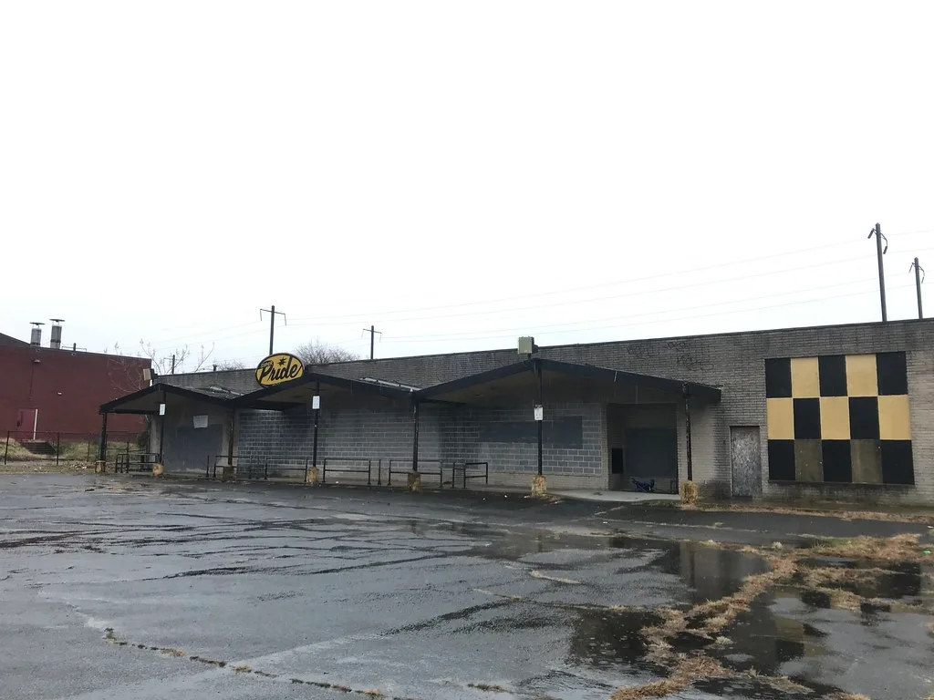 Ancien commerce abandonné à façade grise condamnée sous un auvent à trois pignons, devant un parking fissuré.