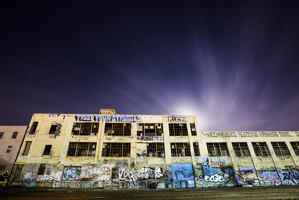 Graffitied old factory with large broken windows photographed at night.