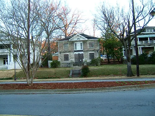 Stone house with boarded openings behind leafless trees.