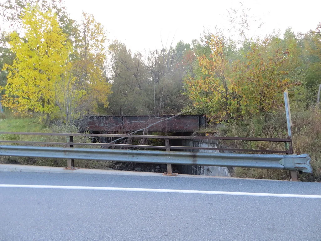 Old rusty metal bridge among trees behind a roadside barrier.