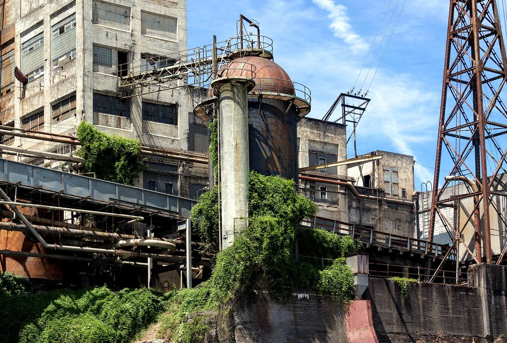 Old factory with broken glazing, a rusty domed tank and ivy-covered pipes.