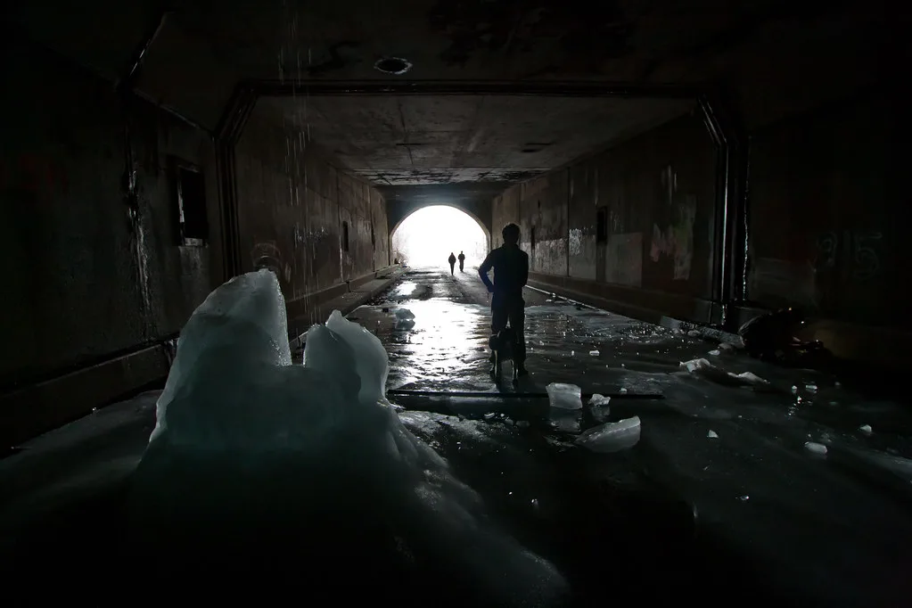 Abandoned passage with damaged walls and blocks of ice on the floor, with silhouettes near the arched exit.
