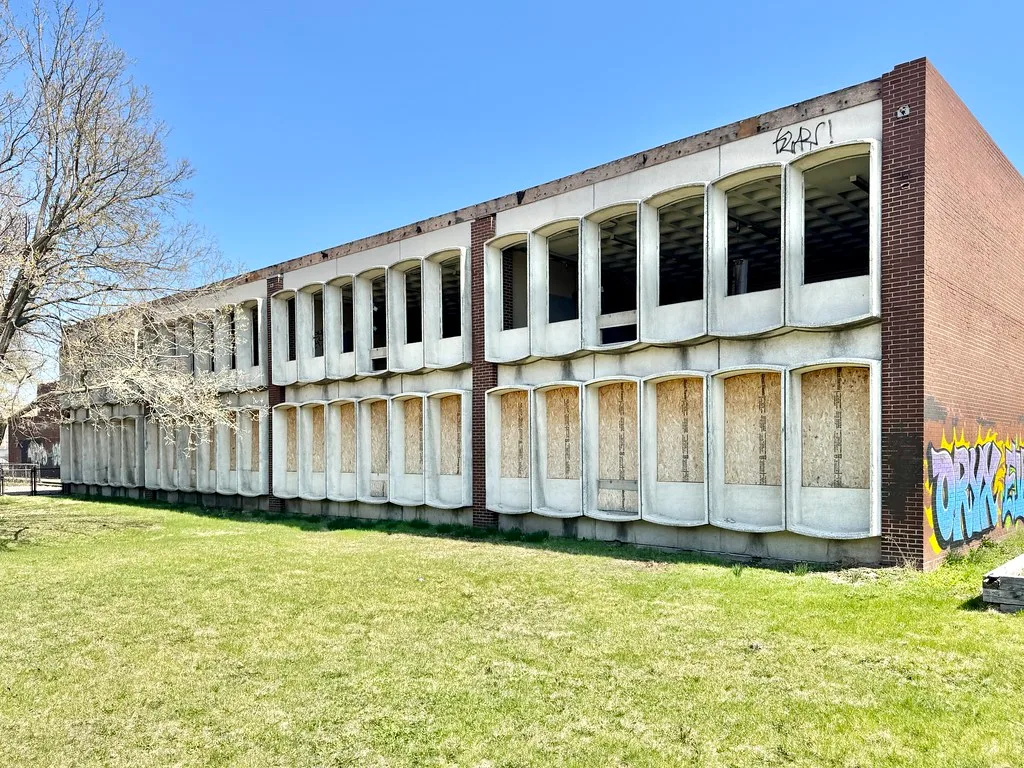 Long abandoned two-story building with a white facade, empty upper windows and wood-covered lower openings.