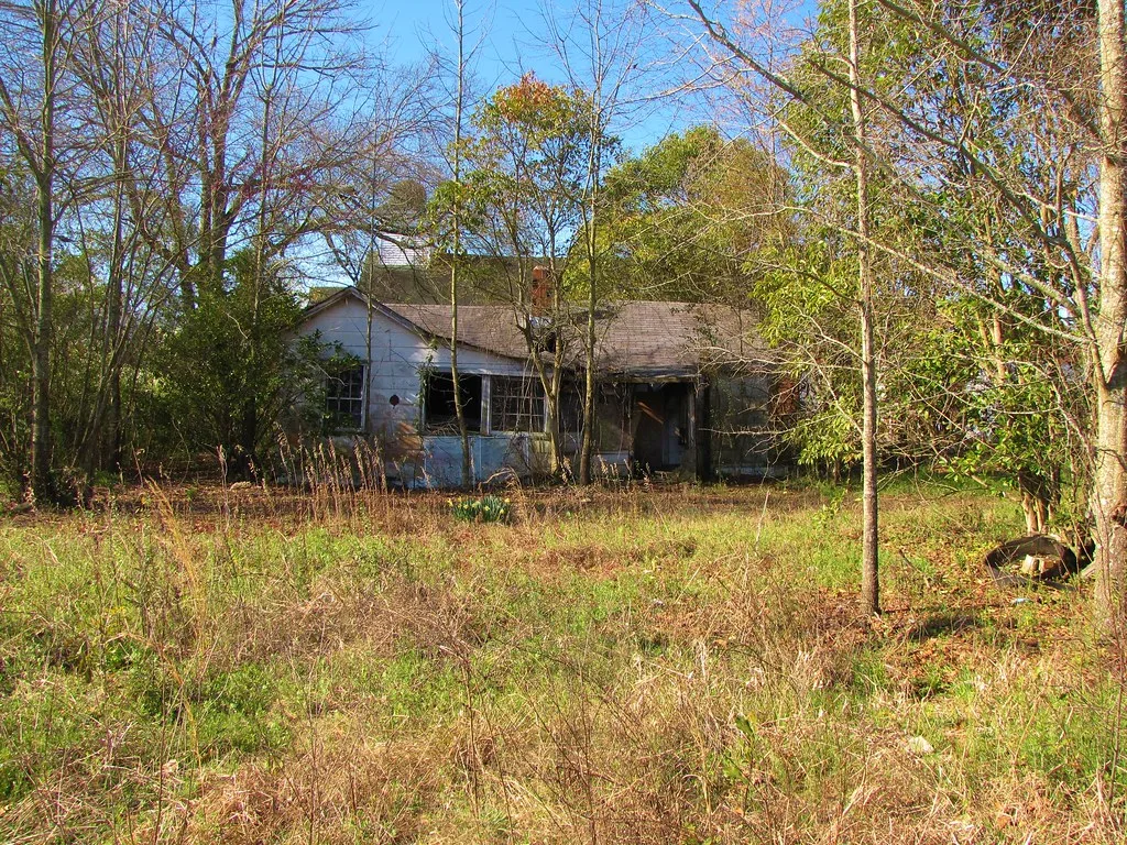 Pale abandoned house with broken windows and a deteriorated dark porch, surrounded by trees and tall grass.