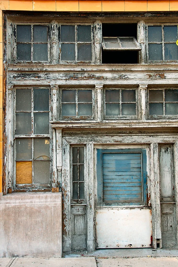 Abandoned shopfront in peeling white wood with partly empty transom windows, a pane replaced by wood and a boarded door with a blue panel.