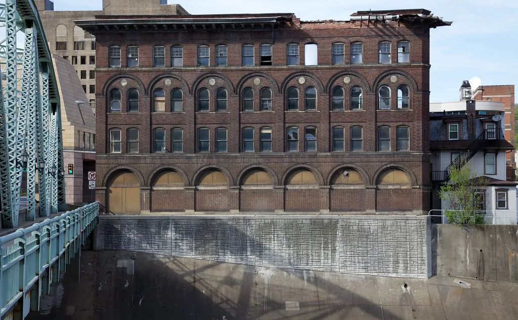 Abandoned brick building with arched windows and a destroyed cornice beside a steel bridge.