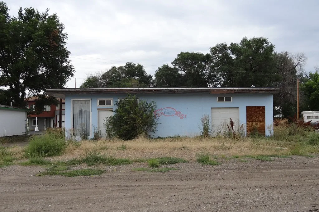 Old pale blue building with boarded doors and overgrown surroundings.