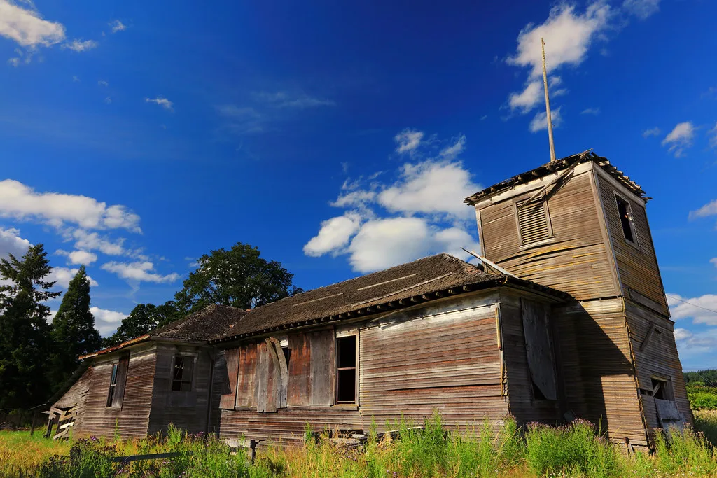 Old wooden building with a deteriorated tower amid tall grass.