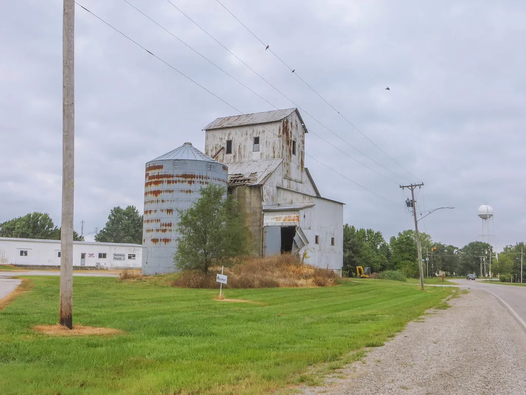 Derelict old white grain elevator with a rusty silo and scrub.