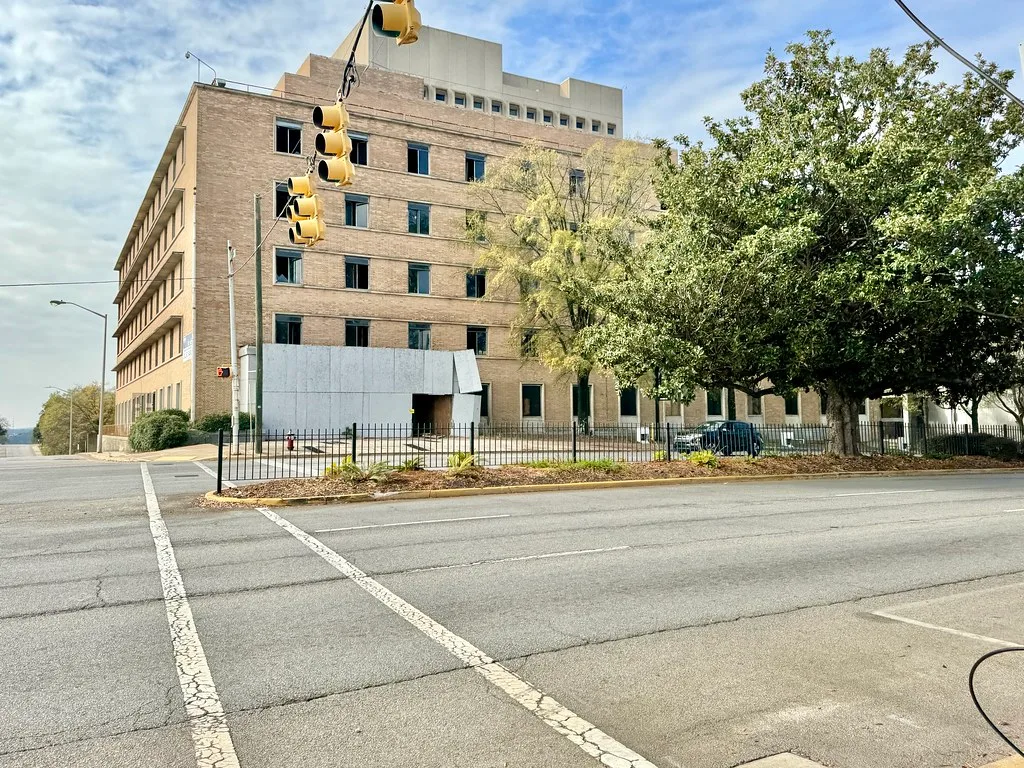 Beige brick building with broken windows and a damaged metal entrance canopy facing a junction.