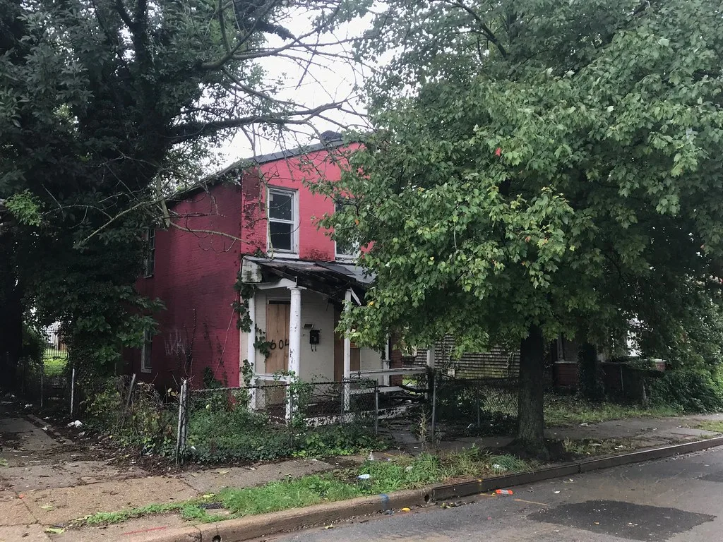 Maison abandonnée rouge à porte condamnée, toiture de porche effondrée et végétation dense derrière une clôture.