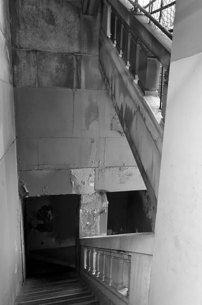 Cage d’escalier abandonnée aux balustrades de pierre et murs écaillés, en noir et blanc.