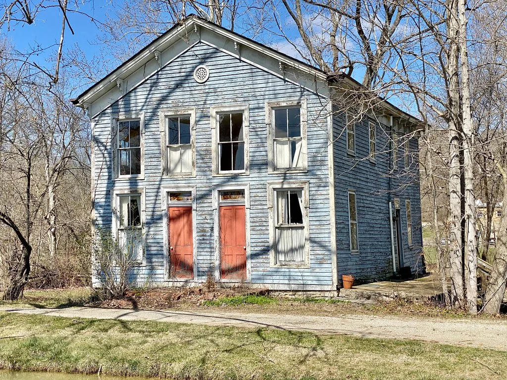 Maison de bois bleu pâle aux deux portes rouges et aux fenêtres cassées parmi les arbres.