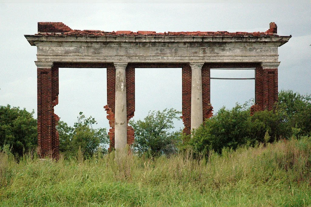 Vestige de façade en brique et pierre à trois grandes ouvertures dans les herbes hautes.