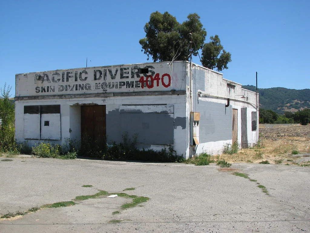 Small white building with a faded sign, boarded doors and weeds along its walls.