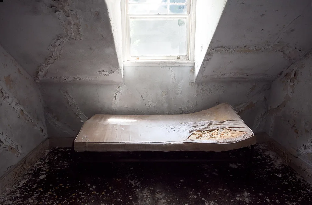 Torn mattress beneath a window in an abandoned room with peeling walls.