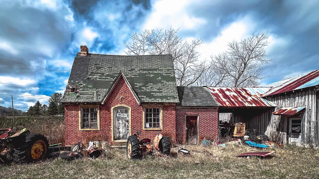 Small red brick building with a holed roof, surrounded by rusty farm machinery and tall grass.