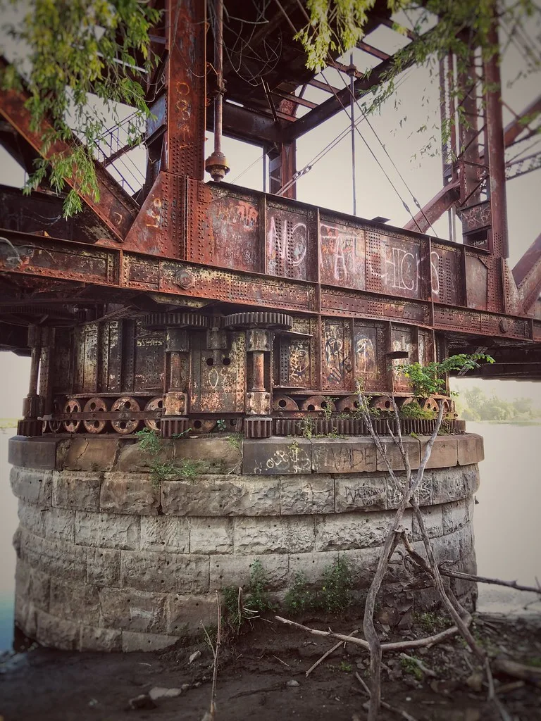 Stone pivot and rusty gears of an old metal bridge overgrown by branches.