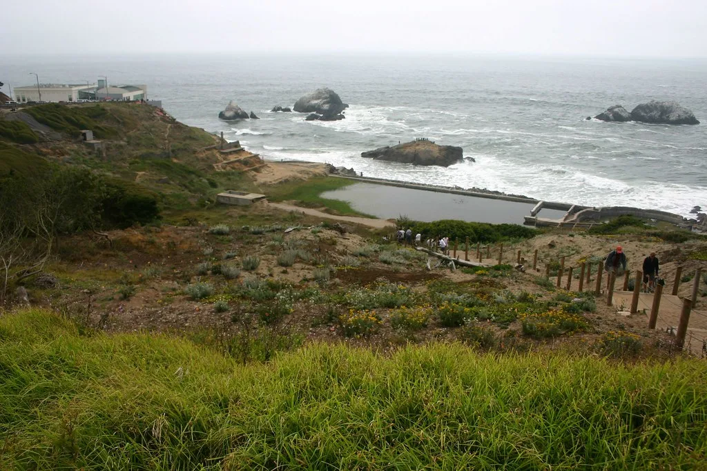 Remains of concrete pools beside the ocean, viewed from a path.