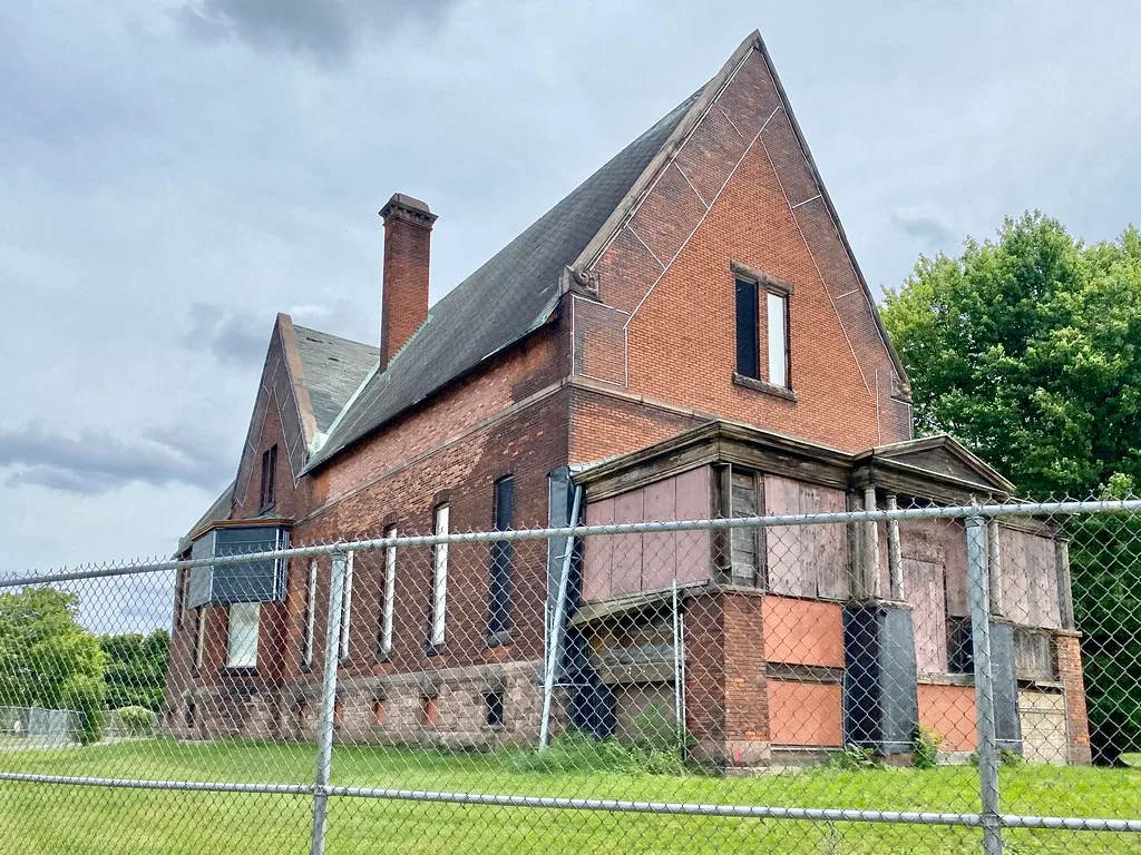Old red-brick building with boarded openings behind a fence.