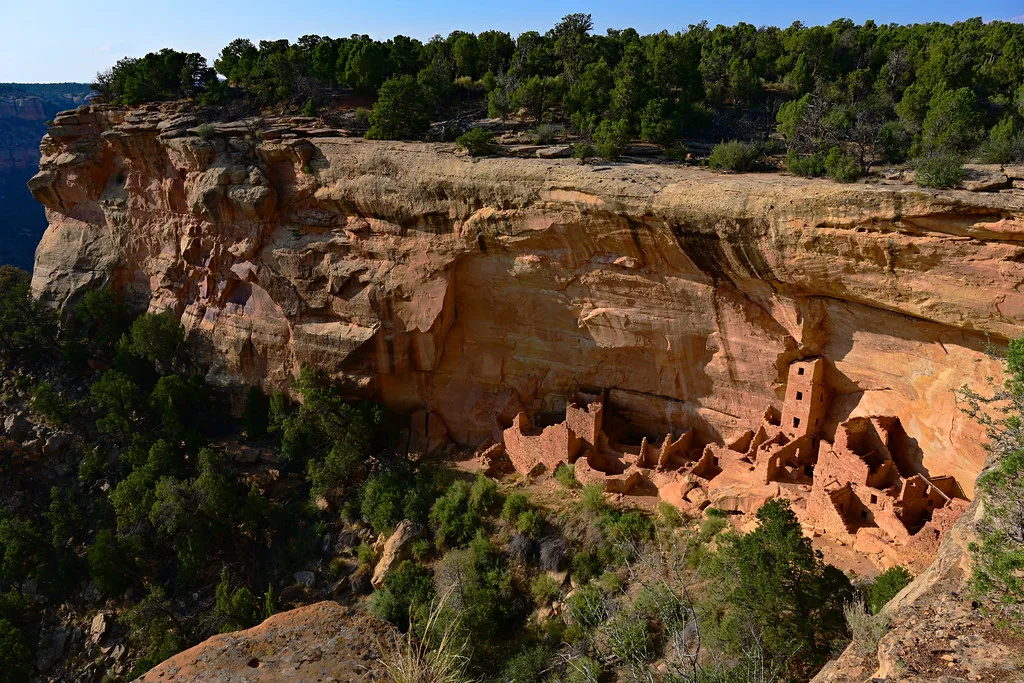 Abandoned ruins of small dwellings and a tower beneath an ochre cliff bordered by trees.