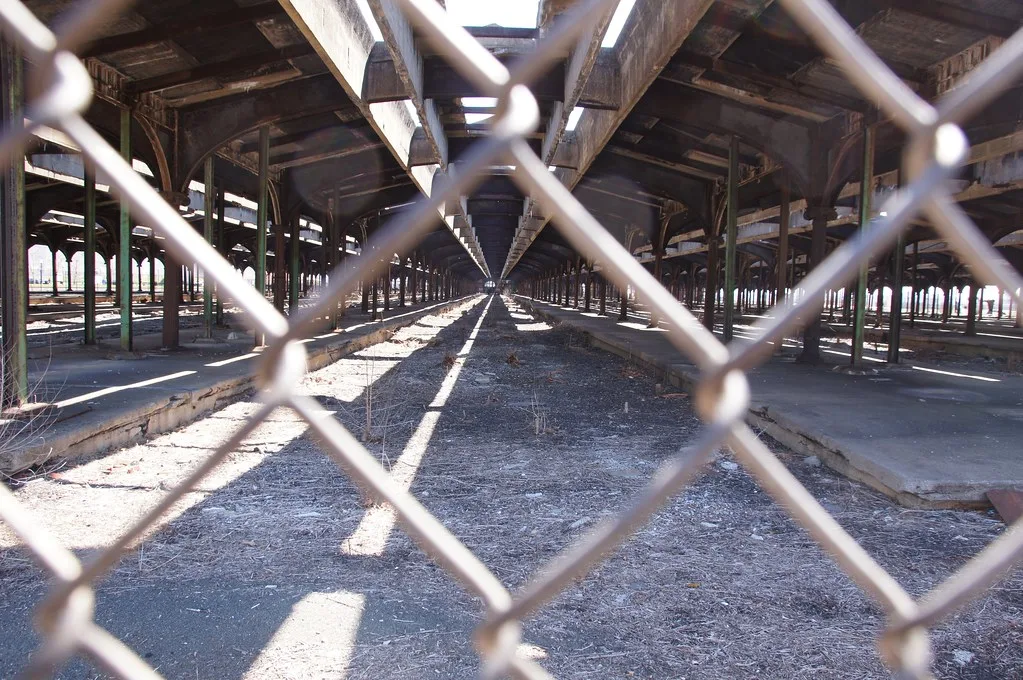 Longue halle ferroviaire abandonnée sans rails, aux quais dégradés et à la charpente rouillée derrière un grillage.