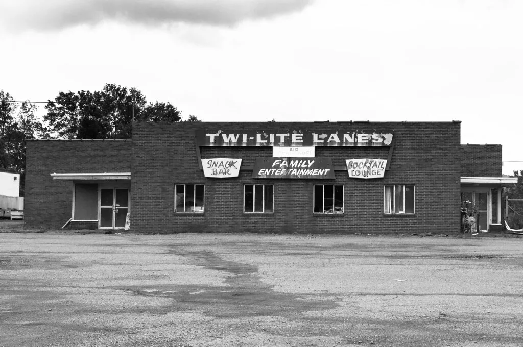 Abandoned former bowling alley with broken windows and a peeling Twi-Lite Lanes sign facing an empty car park, in black and white.