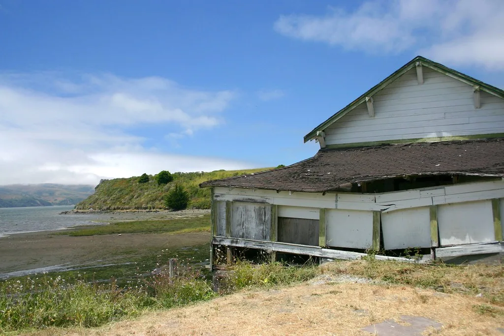 Abandoned white wooden building with a sagging roof and boarded bays, with an exposed base beside a bay.