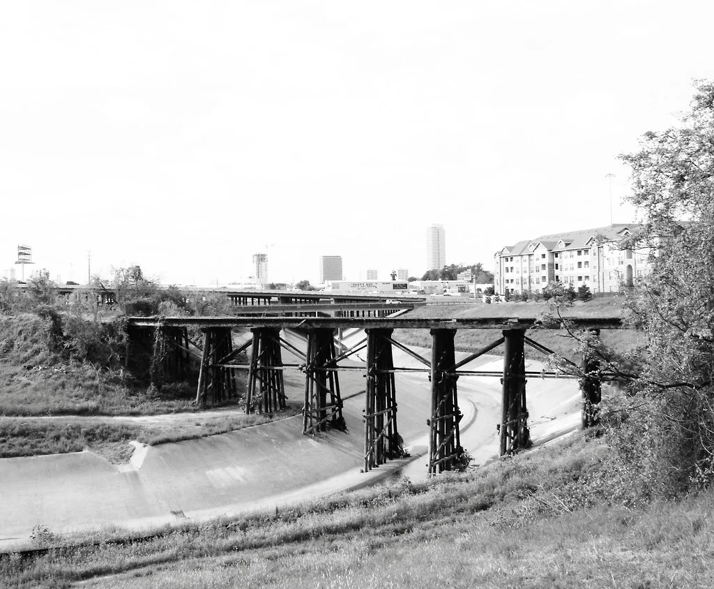 Remains of a timber-pile bridge above a concrete channel with vegetation along its approaches.