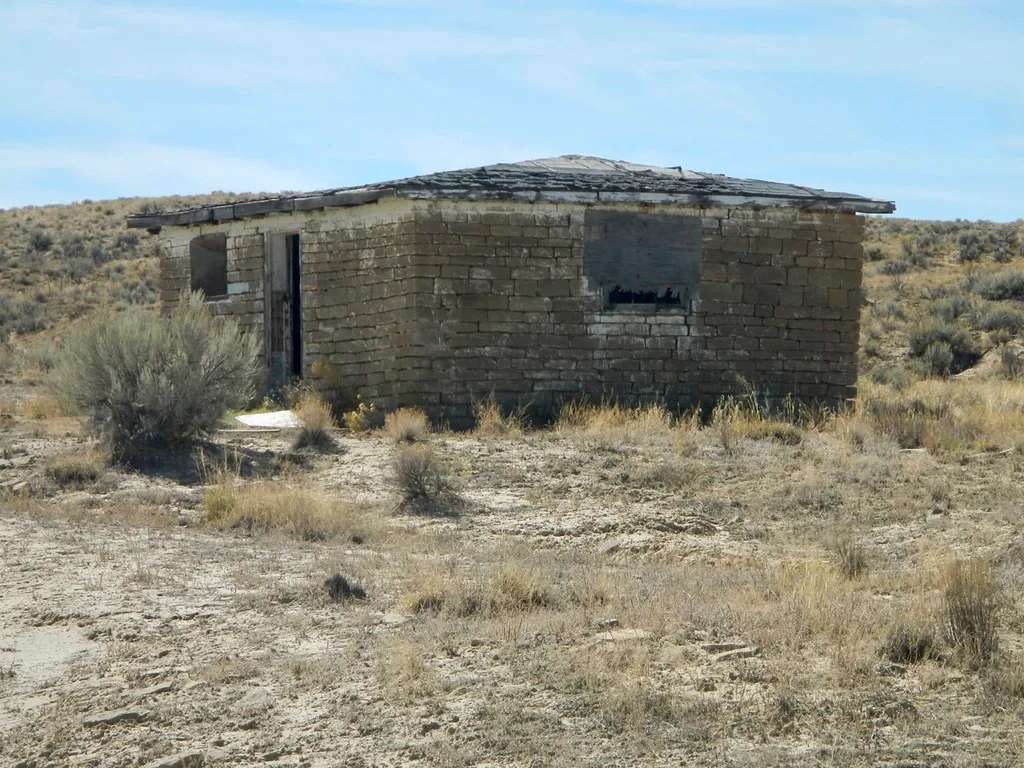 Small abandoned earth-block house with empty windows in a dry shrub-covered landscape.