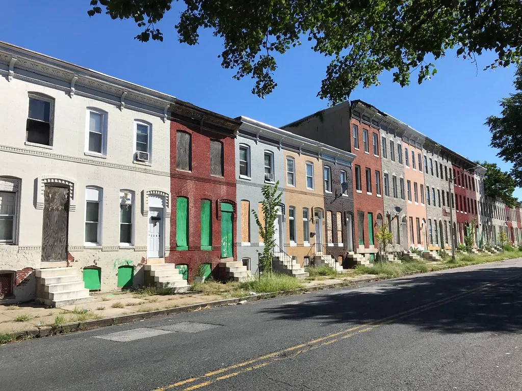 Longue rangée de maisons colorées aux fenêtres couvertes de panneaux et aux entrées envahies d’herbes.
