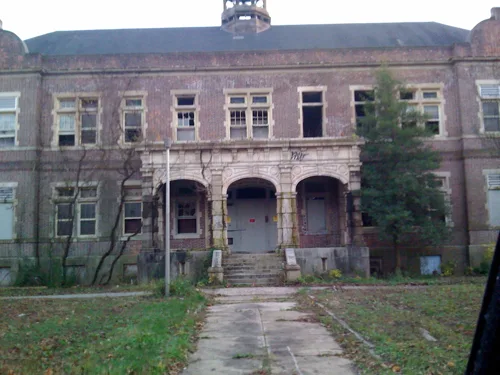 Abandoned old brick building with broken windows and a three-arched porch facing a cracked path.