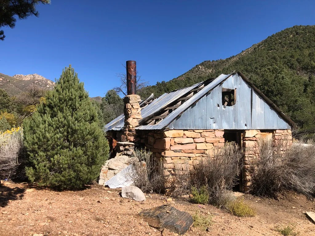 Cabane abandonnée en pierre ocre au toit de tôle troué et cheminée dégradée parmi les buissons d’un versant sec.