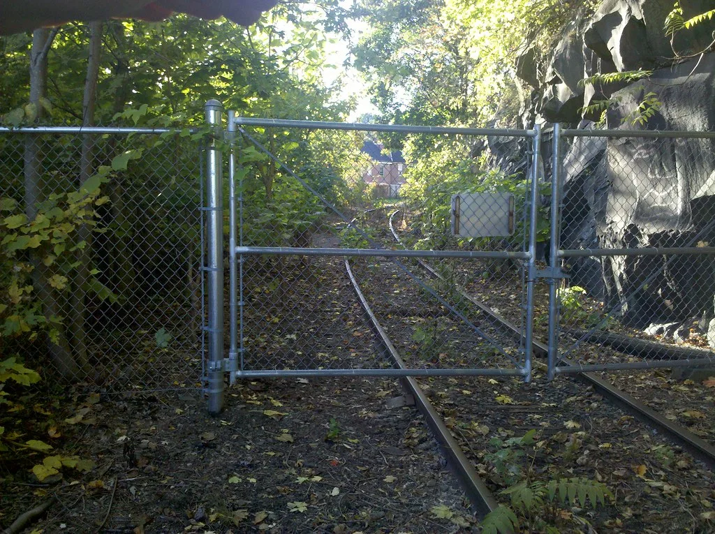 Overgrown railway track behind a chain-link gate.
