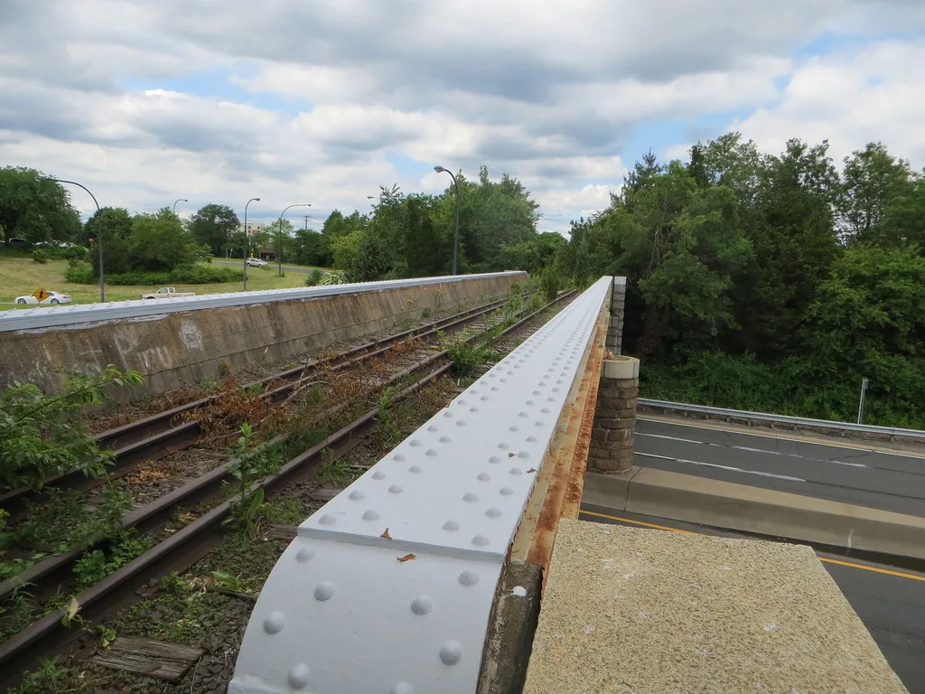 Rails rouillés et traverses envahies de végétation sur un pont métallique au-dessus d’une route.