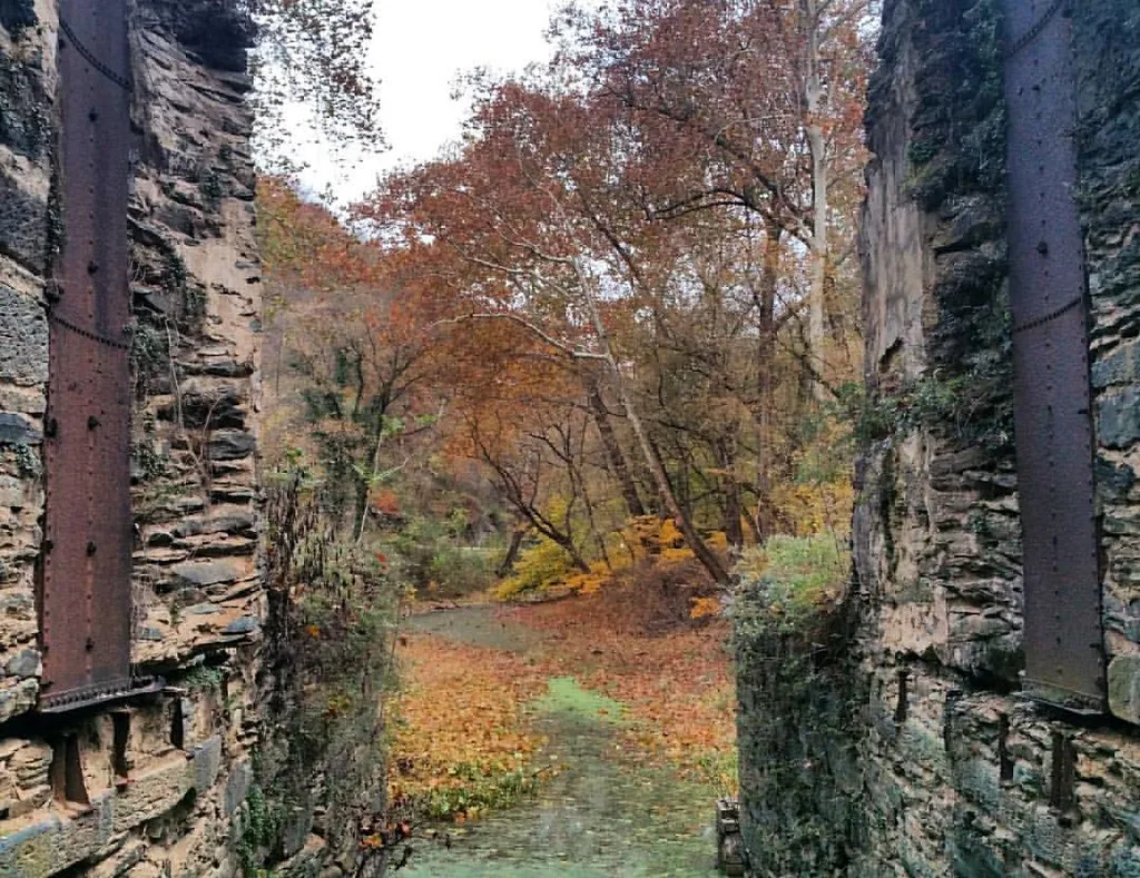 Passage through roofless stone ruins with rusty metal reinforcements, opening onto trees with autumn leaves.