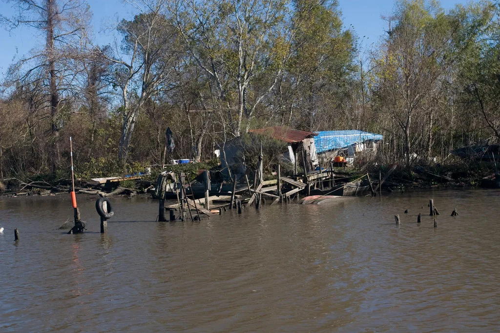 Cabanon au toit partiellement bâché derrière un ponton de bois effondré dans une eau brune.