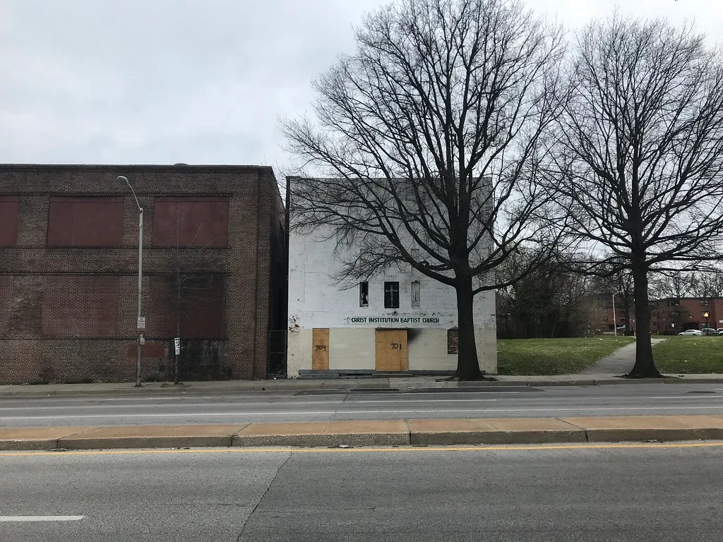 Small white facade with boarded entrances beside a brick building with large sealed windows.