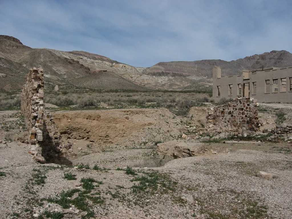 Remains of stone walls and foundations before a large roofless building in the desert.