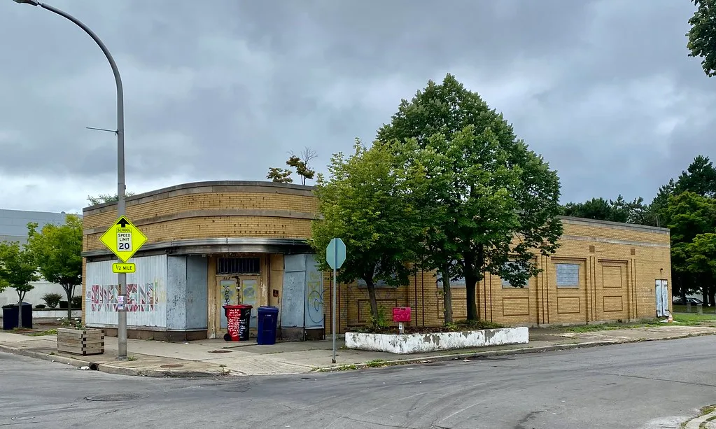 Yellow brick corner commercial building with sealed shop windows and graffitied panels.