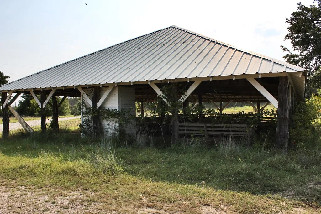 Old metal-roofed pavilion overgrown with plants and tall grass.