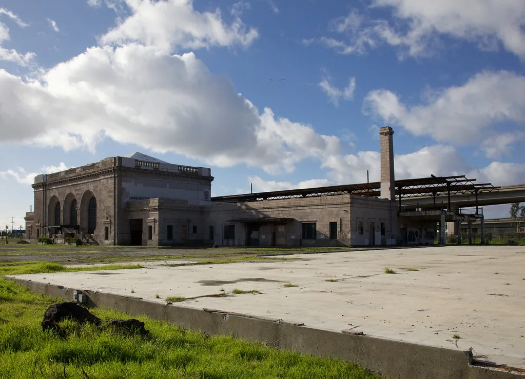 Abandoned former station with tall arched windows and dismantled platform canopies behind a cracked concrete platform.