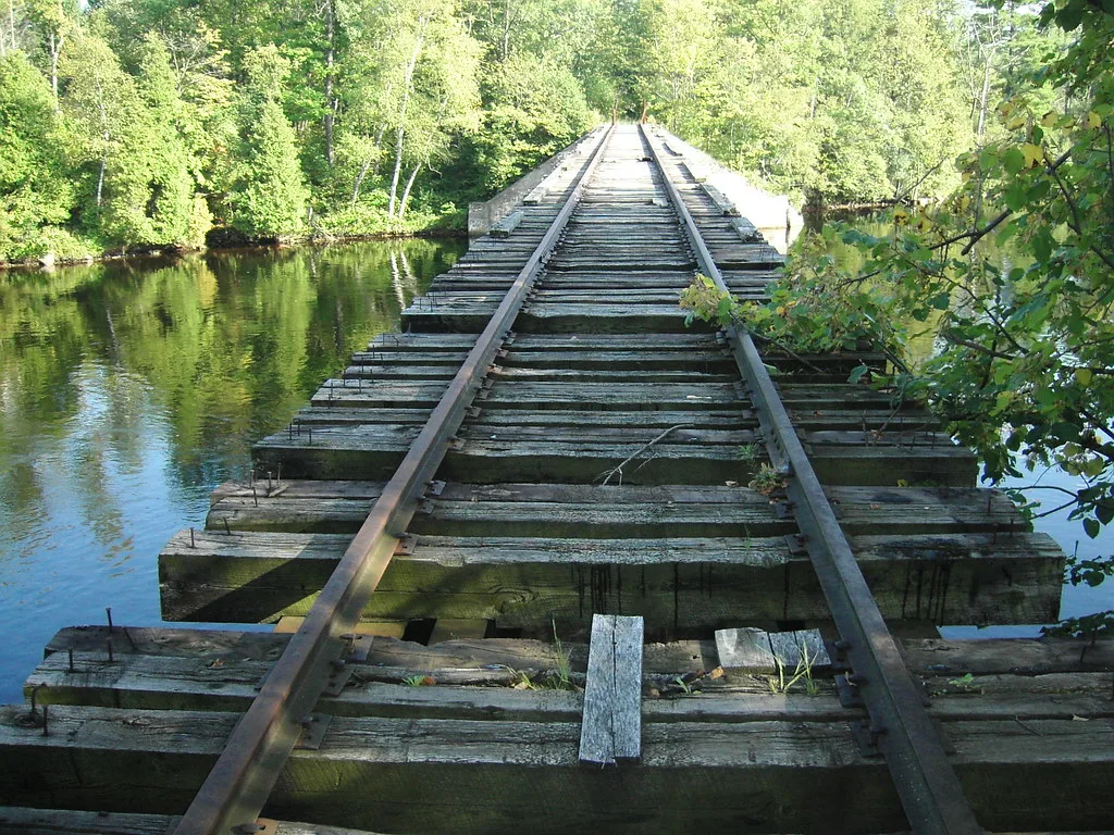 Abandoned railway bridge over a river, with split and displaced wooden sleepers and rusty rails among trees.