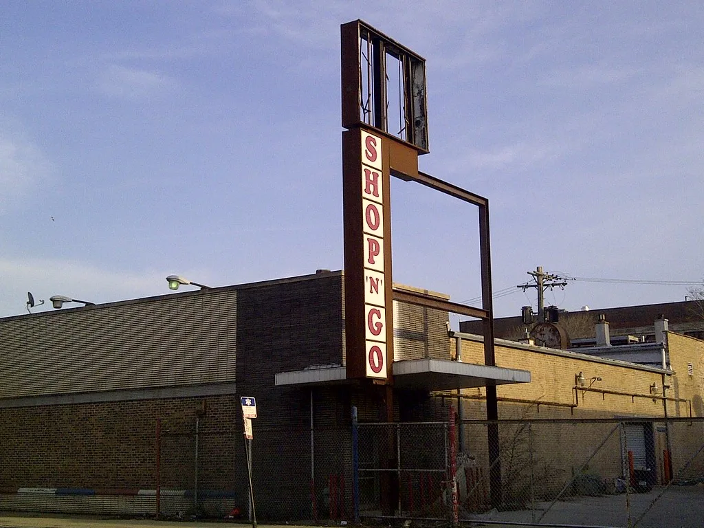 Disused former shop behind a wire fence with a large partly dismantled vertical sign.